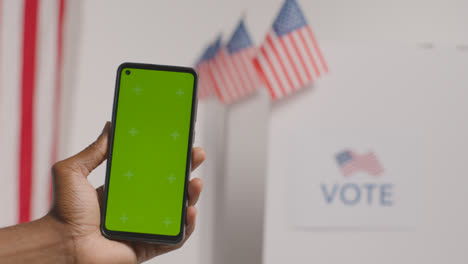 Nahaufnahme-Einer-Hand,-Die-Ein-Mobiltelefon-Mit-Grünem-Bildschirm-Vor-Der-Wahlurne-Bei-Der-Amerikanischen-Wahl-1-Hält